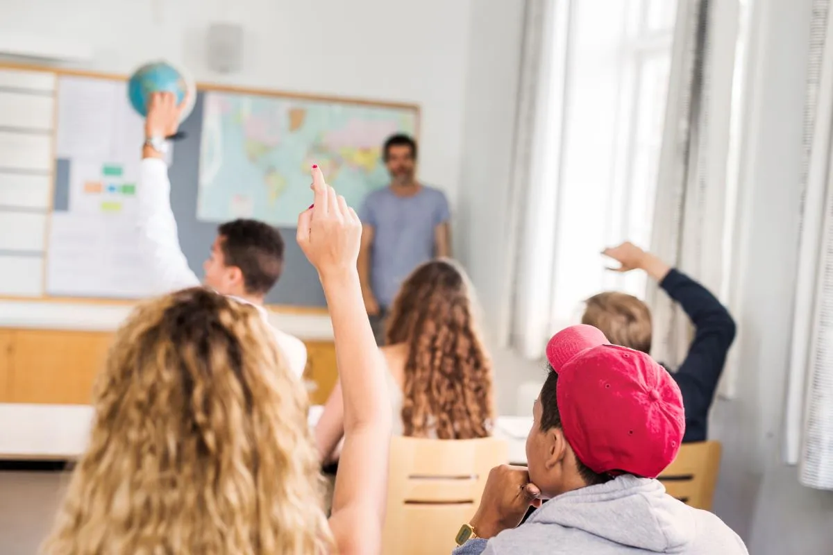 supply teacher in classroom in cambridge full of pupils, some with raised arms